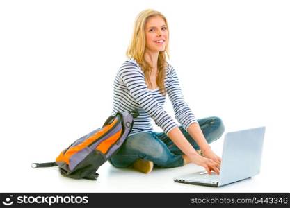 Smiling teengirl sitting on floor with schoolbag and using laptop isolated on white &#xA;