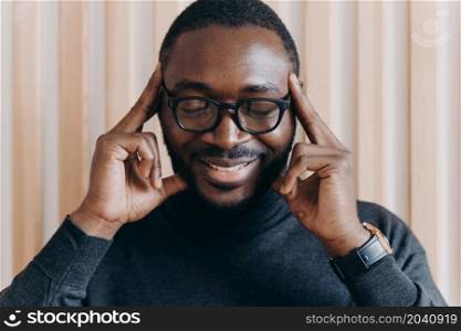 Smiling thoughtful African American male entrepreneur posing with closed eyes and with forefingers touching temples, showing brooding expression and birth of new ideas and successful projects. Thoughtful African American male entrepreneur with closed eyes touching temples with forefingers