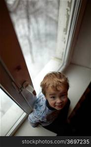 Smiling two year old boy sits on the windowsill and looking to the camera.
