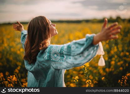 Smiling ukrainian woman standing in yellow canola field. Young lady in blue embroidery vyshyvanka blouse. Ukraine, independence, freedom, patriot symbol, victory in war.. Happy free ukrainian woman in embroidery blouse in yellow canola field.Open arms