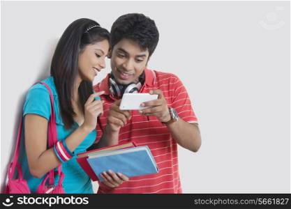 Smiling university friends reading text message on smart phone against wall