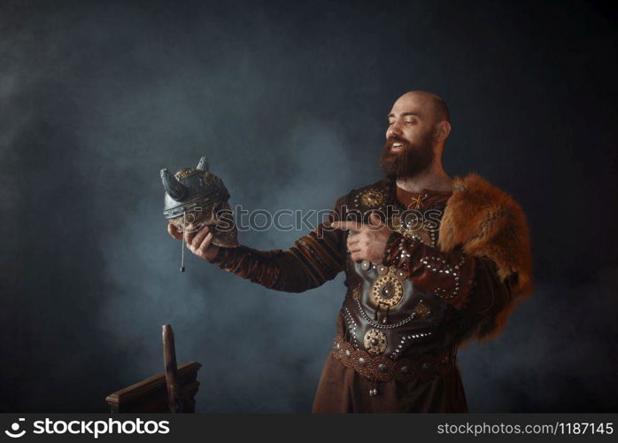 Smiling viking dressed in traditional nordic clothes holds enemy&rsquo;s skull in helmet, barbarian image. Ancient warrior in smoke on dark background