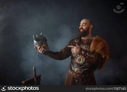 Smiling viking dressed in traditional nordic clothes holds enemy&rsquo;s skull in helmet, barbarian image. Ancient warrior in smoke on dark background