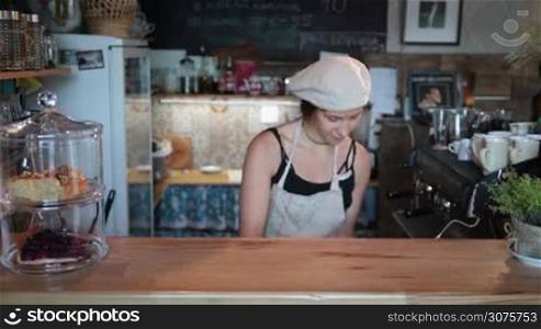 Smiling waitress in apron putting on counter a tasty cheesecake on cake stand . Attractive female barista openning a cake stand with delicious cheesecake and giving thumb up