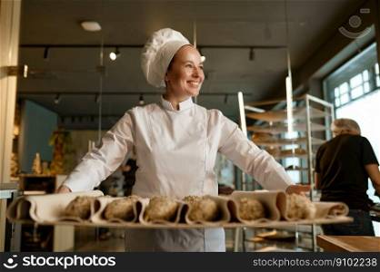 Smiling woman baker preparing sweet buns pastries with sesame seeds while working at professional kitchen of modern bakery house. Smiling baker preparing sweet buns pastries with sesame seeds