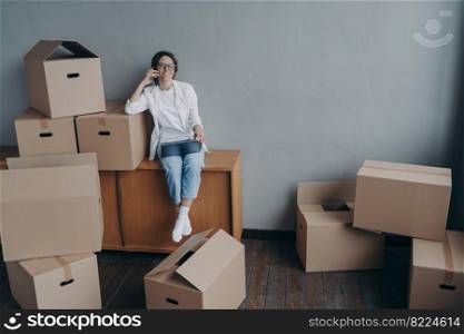 Smiling woman calling moving company, ordering mover for relocation to new house, sitting with laptop surrounded cardboard boxes. Real estate rental, mortgage, purchase of housing concept.. Female calling moving company, orders mover for relocation, sitting with laptop and cardboard boxes