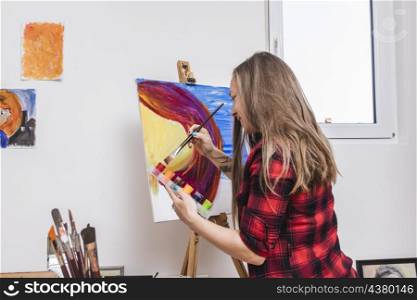 smiling woman dipping paintbrush into gouache jar standing by easel