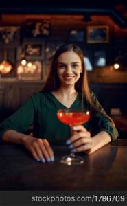 Smiling woman drinks coctail at the counter in bar. One female person in pub, human emotions, leisure activities, night life. Smiling woman drinks coctail at the counter in bar