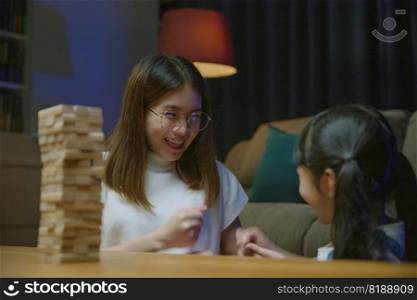 Smiling woman help teach child play build constructor of wooden blocks, Asian young mother playing game in wood block with her little daughter in home living room at night time before going to bed