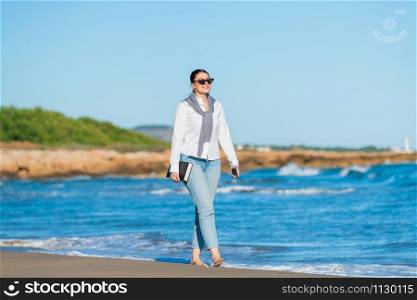 Smiling woman holding a book and her phone walks along the beach on a sunny day on an out of focus background. Leisure concept.. Smiling woman walking along the sunny beach