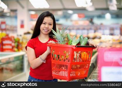 Smiling woman holding basket filled with fruits in shopping centre and looking at camera