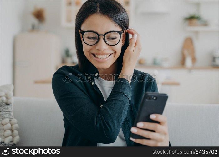 Smiling woman holding smartphone reading message chatting in social networks, sitting on couch at home. Portrait of excited girl wearing glasses received good news, got promotion at work.. Excited woman holding smartphone got good news message in social networks, sitting on couch at home