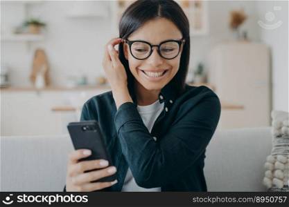 Smiling woman holding smartphone reading message chatting in social networks, sitting on couch at home. Portrait of excited girl wearing glasses received good news, got promotion at work.. Excited woman holding smartphone got good news message in social networks, sitting on couch at home
