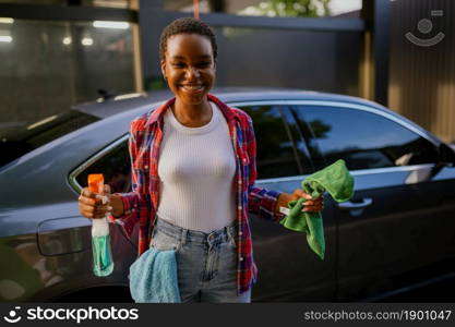 Smiling woman holds window cleaner spray and a rag, hand car wash station. Car-wash industry or business. Female person cleans her vehicle from dirt outdoors. Woman holds window cleaner and rag, hand car wash