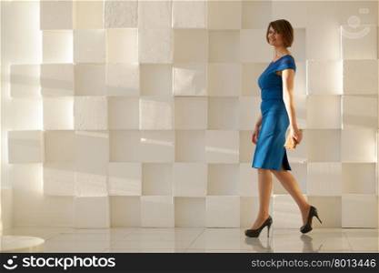 Smiling woman in elegant blue dress walking against modern wall with wallet in hand while looking at camera. Smiling woman in blue dress walking against modern wall with wallet in hand