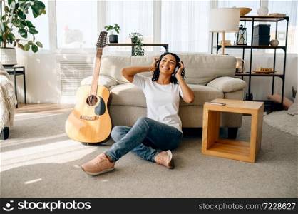 Smiling woman in headphones at home, guitar on background. Pretty lady with musical instrument relax in the room, female music lover resting. Smiling woman in headphones, guitar on background
