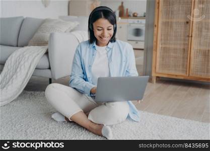 Smiling woman in headphones working on laptop from home learning online sitting on warm floor. Friendly female student in wireless headset listening webinar. Distance education, e-learning.. Smiling woman in headset working on laptop from home learning online sitting on the floor. Elearning