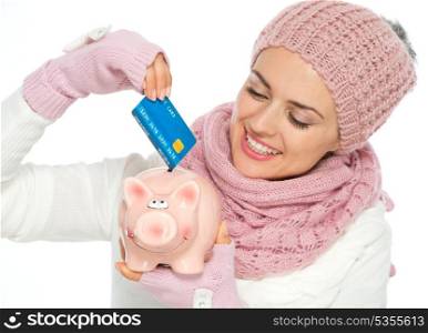 Smiling woman in knit scarf, hat and mittens putting credit card in piggy bank