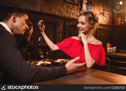 Smiling woman in red dress makes image of her man on phone camera. Beautiful love couple in restaurant, romantic evening, anniversary celebration. Woman in red dress makes image of her man on phone