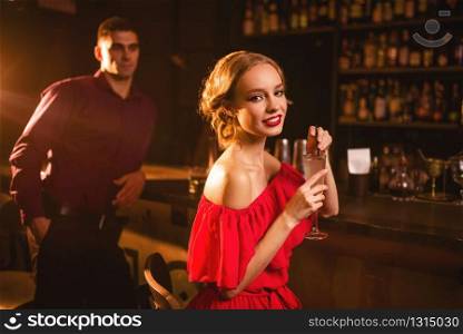 Smiling woman in red dress with cocktail in hand, man behind bar counter, flirting. Date in nightclub, attractive love couple in pub