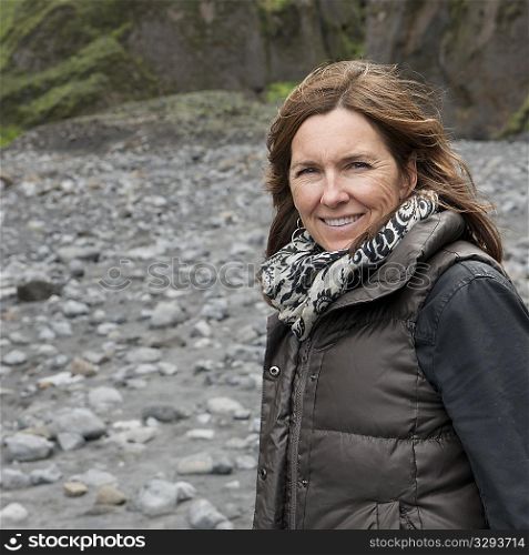 Smiling woman in scarf and vest at rocky dry riverbed before cliff