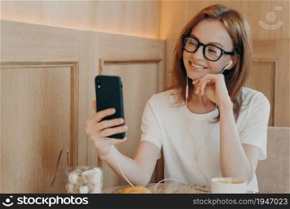Smiling woman in spectacles wears earphones uses smartphone in cafe, young positive female sits at table in coffee shop, has coffee and enjoys video chat call with family or friend on mobile phone. Smiling happy woman in spectacles wears earphones uses smartphone in cafe