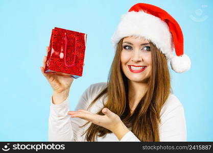 Smiling woman is happy to give Christmas gifts. Female wearing santa claus hat holding present red gift bag, on blue. Happy Christmas woman holds red gift bag