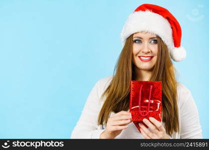 Smiling woman is happy to give Christmas gifts. Female wearing santa claus hat holding present red gift bag, on blue. Happy Christmas woman holds red gift bag