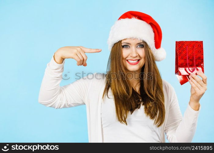 Smiling woman is happy to give Christmas gifts. Female wearing santa claus hat points to present red gift bag, on blue. Christmas woman points to red gift bag