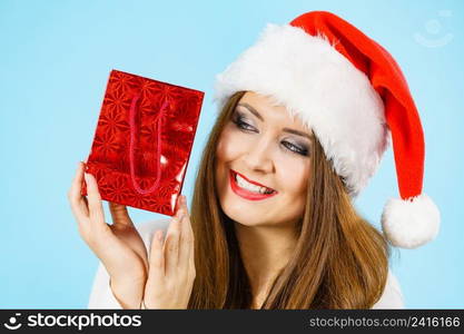 Smiling woman is happy to give Christmas gifts. Female wearing santa claus hat holding present red gift bag, on blue. Happy Christmas woman holds red gift bag