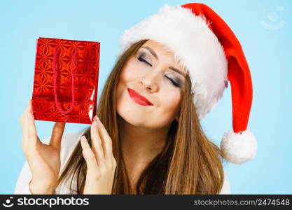 Smiling woman is happy to give Christmas gifts. Female wearing santa claus hat holding present red gift bag, on blue. Happy Christmas woman holds red gift bag