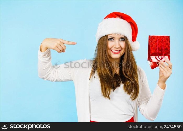 Smiling woman is happy to give Christmas gifts. Female wearing santa claus hat points to present red gift bag, on blue. Christmas woman points to red gift bag
