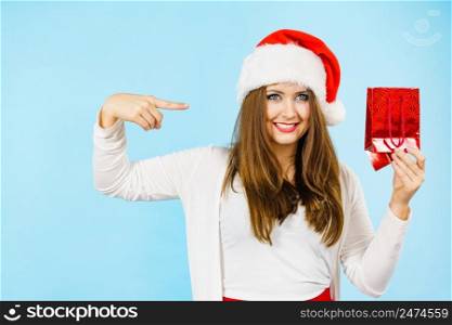 Smiling woman is happy to give Christmas gifts. Female wearing santa claus hat points to present red gift bag, on blue. Christmas woman points to red gift bag