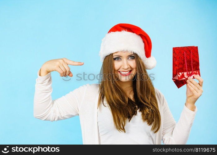 Smiling woman is happy to give Christmas gifts. Female wearing santa claus hat points to present red gift bag, on blue. Christmas woman points to red gift bag