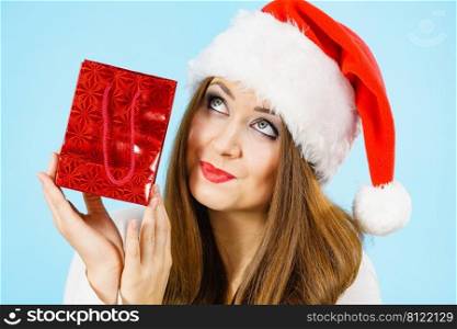 Smiling woman is happy to give Christmas gifts. Female wearing santa claus hat holding present red gift bag, on blue. Happy Christmas woman holds red gift bag