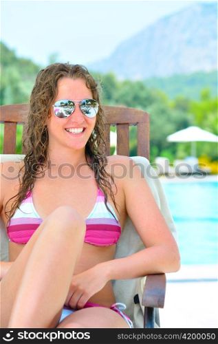 smiling woman is sitting in chaise longue near a swimming pool