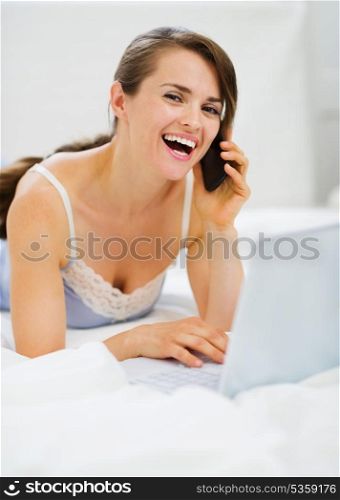 Smiling woman laying on bed with laptop making phone call