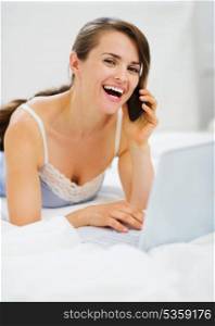 Smiling woman laying on bed with laptop making phone call