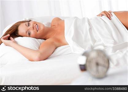 Smiling woman lying in white bed, alarm clock in foreground