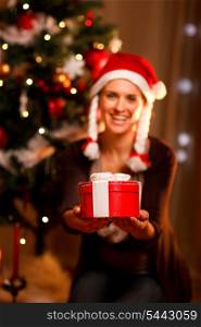 Smiling woman near Christmas tree presenting gift box. Focus on gift&#xA;