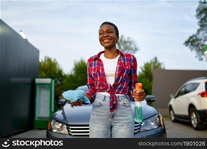 Smiling woman poses with window cleaner spray and a rag, hand car wash station. Car-wash industry or business. Female person cleans her vehicle from dirt outdoors. Woman poses with spray and a rag, car wash station