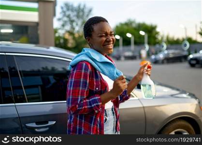 Smiling woman poses with window cleaner spray and a rag, hand car wash station. Car-wash industry or business. Female person cleans her vehicle from dirt outdoors. Woman poses with spray and a rag, car wash station