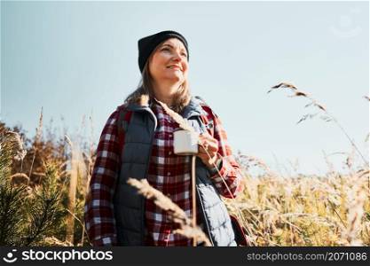 Smiling woman relaxing and enjoying the coffee during summer trip. Woman standing on trail and looking away holding cup of coffee. Woman with backpack hiking through tall grass along path in mountains. Spending summer vacation close to nature