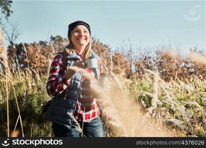 Smiling woman relaxing and enjoying the coffee during summer trip. Woman standing on trail and looking away holding cup of coffee. Woman with backpack hiking through tall grass along path in mountains. Spending summer vacation close to nature
