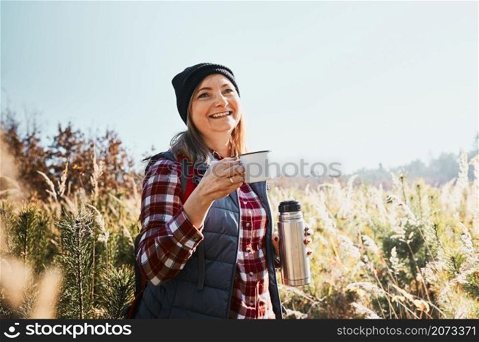Smiling woman relaxing and enjoying the coffee during vacation trip. Woman standing on trail and looking away holding cup of coffee and thermos flask. Woman with backpack hiking through tall grass along path in mountains. Active leisure time close to nature. Smiling woman relaxing and enjoying the coffee during vacation trip. Woman standing on trail and looking away holding cup of coffee and thermos flask. Woman with backpack hiking through tall grass along path in mountains. Active leisure time close to natu