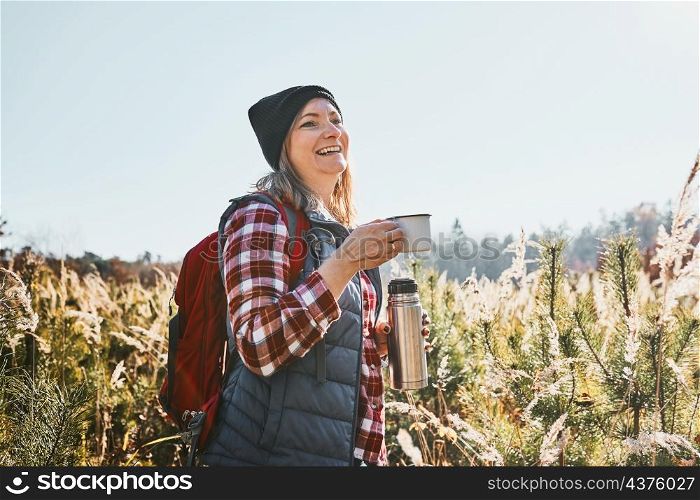 Smiling woman relaxing and enjoying the coffee during vacation trip. Woman standing on trail and looking away holding cup of coffee and thermos flask. Woman with backpack hiking through tall grass along path in mountains. Active leisure time close to nature. Smiling woman relaxing and enjoying the coffee during vacation trip. Woman standing on trail and looking away holding cup of coffee and thermos flask. Woman with backpack hiking through tall grass along path in mountains. Active leisure time close to natu