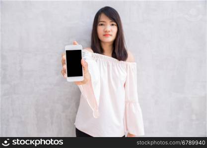 Smiling woman showing a blank smartphone screen standing on concrete wall.