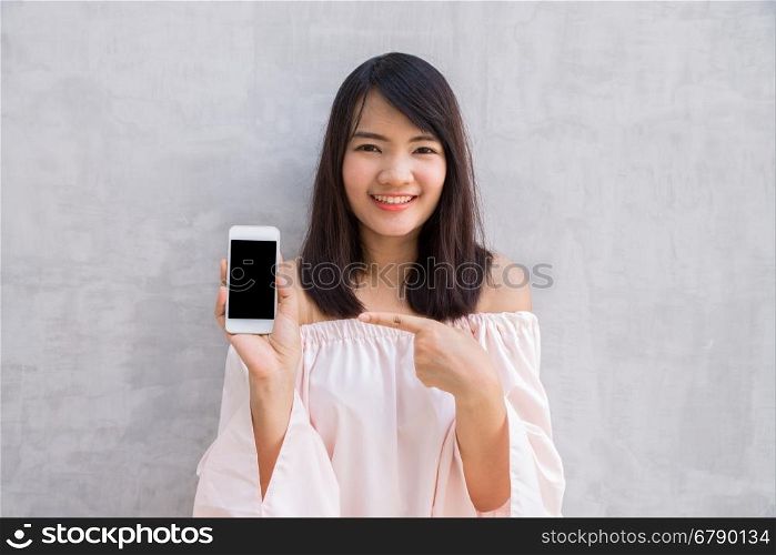 Smiling woman showing a blank smartphone screen standing on concrete wall.
