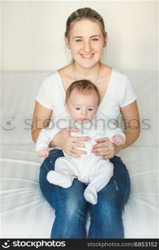 Smiling woman sitting at bedroom and holding her cute baby on laps