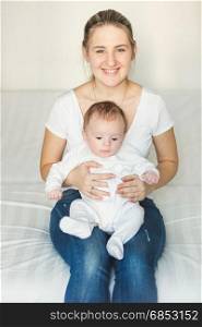 Smiling woman sitting at bedroom and holding her cute baby on laps